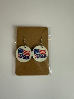 USA flag earrings attached to a brown backing card inside a clear plastic sleeve, showing white circular earrings with “USA!” text and flag motif in red, white, and blue.