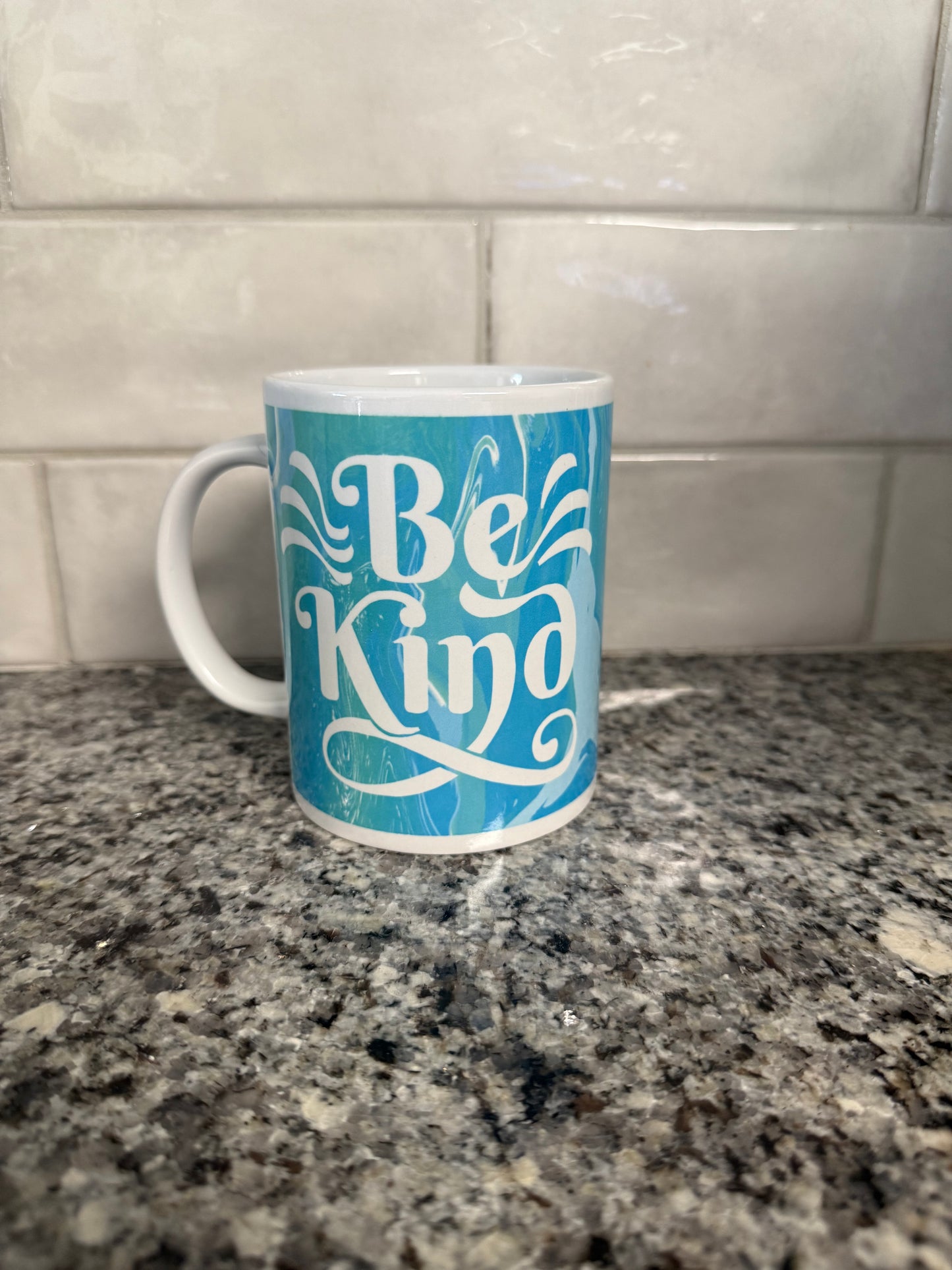Left side view of "Be Kind' ceramic coffee mug in blue and green with white handle Roxzell