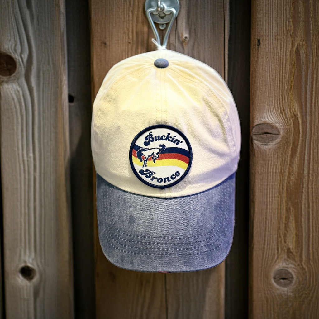 A vintage Buckin' Bronco baseball cap hanging on a metal hook against a rustic wooden plank wall. The cap features a cream-colored crown, a denim visor, and a circular embroidered logo patch on the front. Roxzell