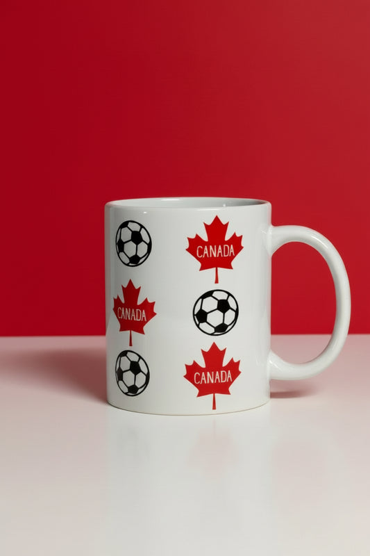 White ceramic mug decorated with red maple leaves labeled “Canada” and black‑and‑white soccer balls on a red and white background, symbolizing Canadian soccer pride. Roxzell