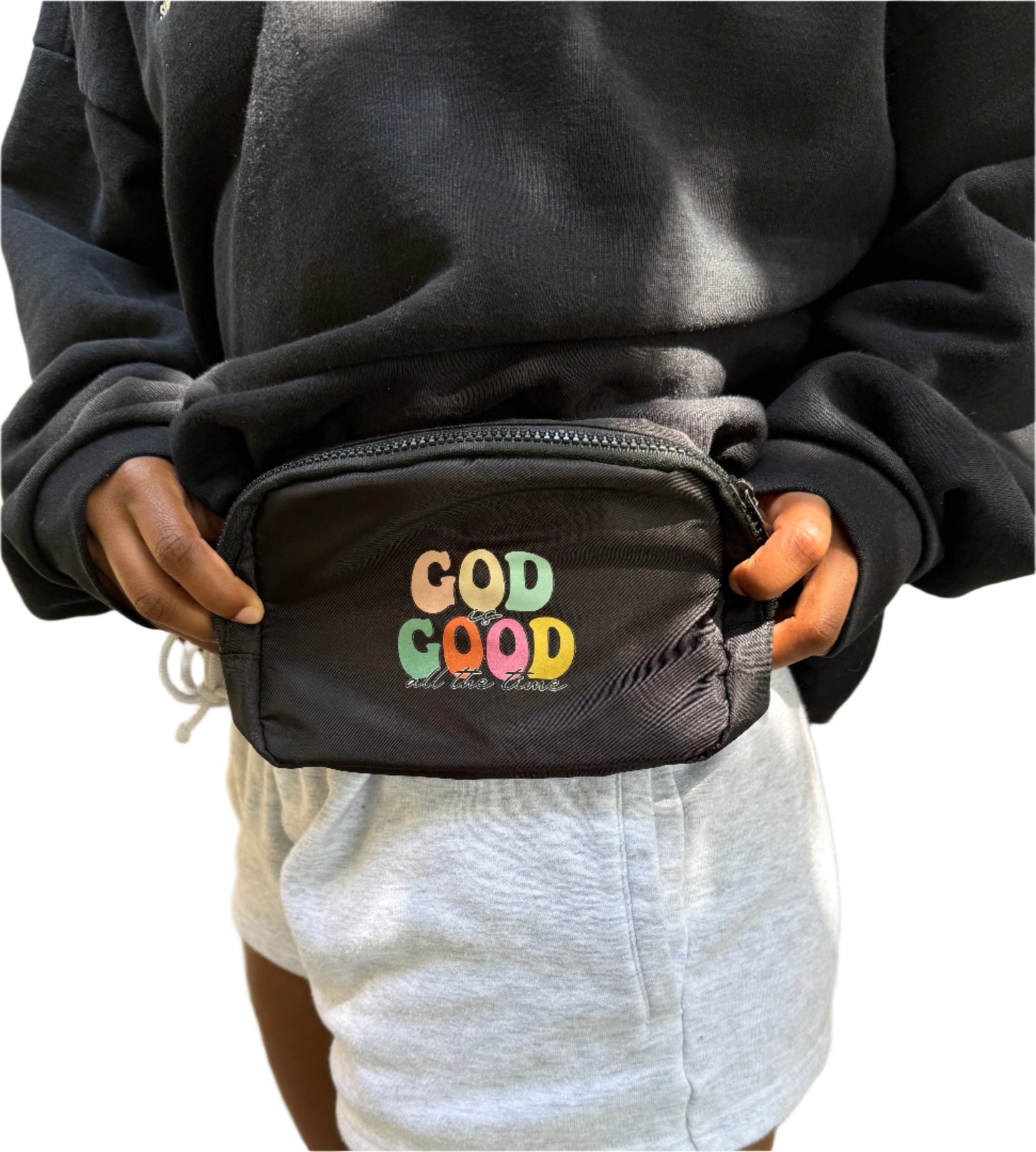 Person holding a black fanny pack/shoulder bag with colorful 'God is Good' text on the front.