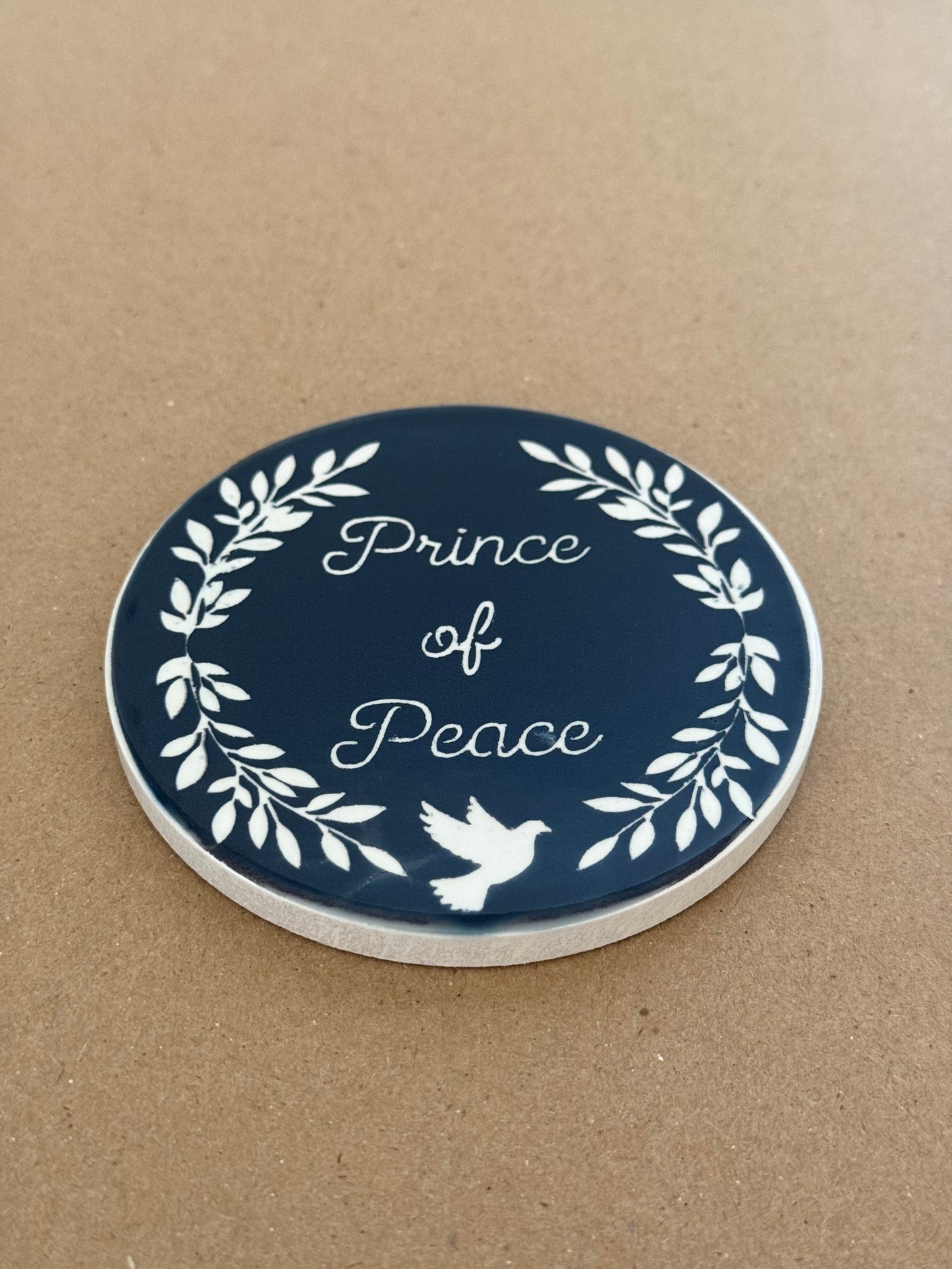 Kitchen table-top drink coaster. Coaster is midnight blue designed with the words "Prince of Peace", leaves and a dove, all in white.