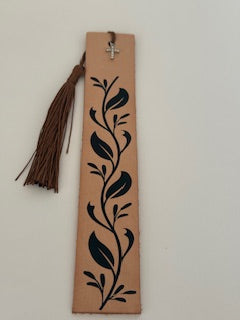 A tan leather bookmark with a black vine pattern, cross charm, and a brown tassel, placed on a neutral background.