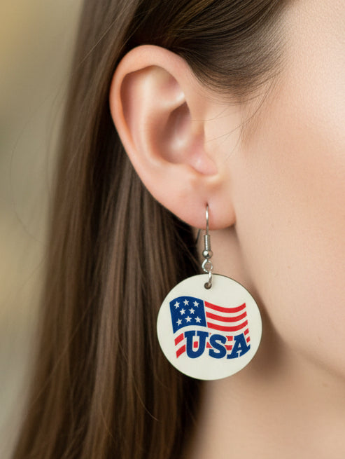 Person wearing a round earrings with metal hooks showing a patriotic design of the American flag and the word “USA” in blue letters. Roxzell