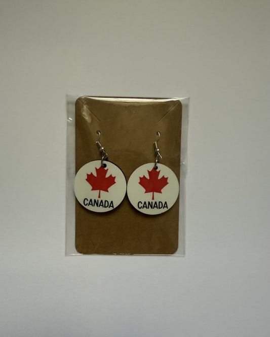 Canada maple leaf earrings attached to a brown backing card inside a clear plastic sleeve, showing red maple leaf designs with black “Canada” text below the leaf. Roxzell