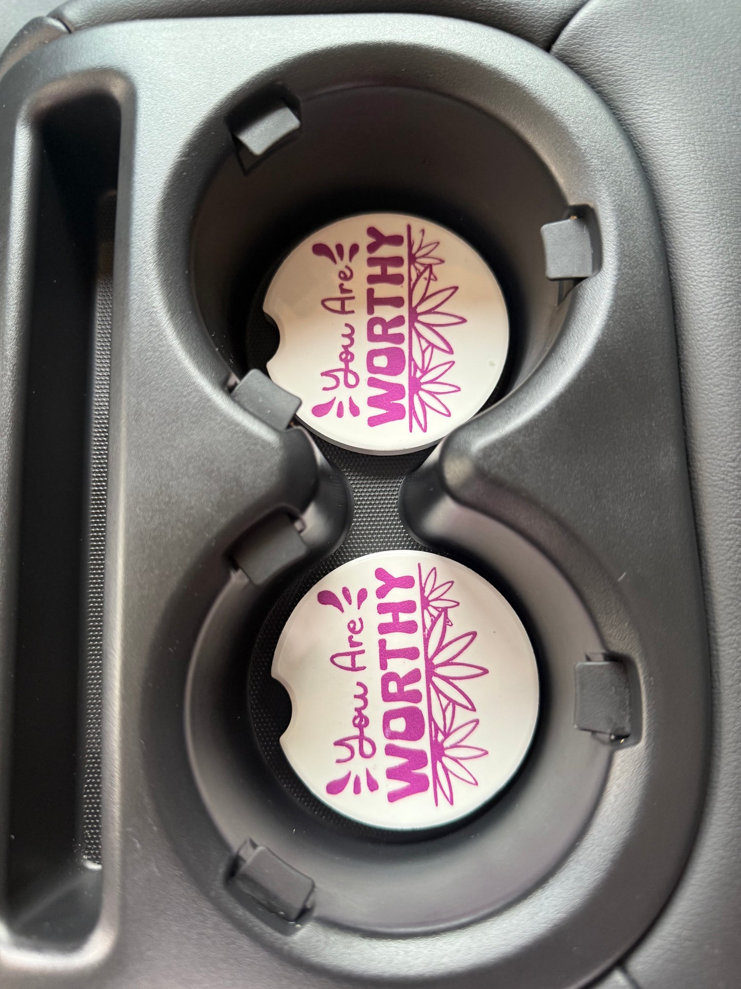 Two car cup holders with 'You Are' Worthy" car coasters inside a vehicle.