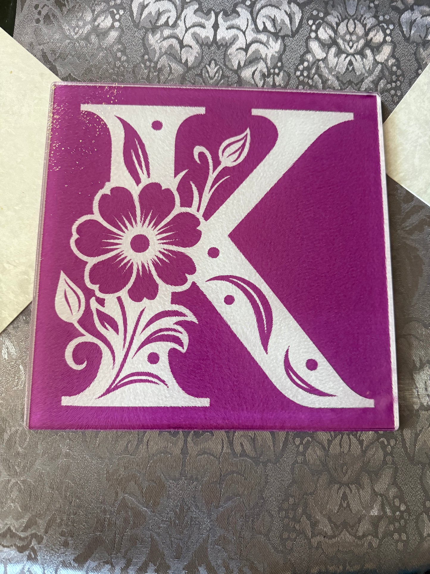 Tempered glass board with intricate white floral "K" design, placed on textured fabric table runner, Roxzell