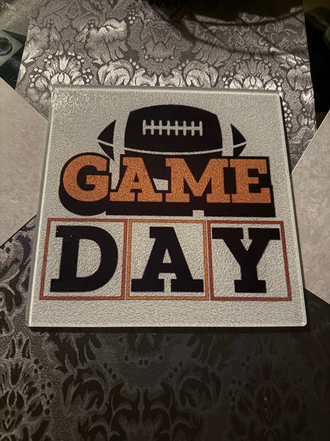 Acrylic glass cutting board with a brown football and orange and brown "Game Day" theme on a table with a gray pattern table runner.