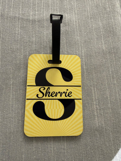 Personalized yellow luggage tab with the name Sherrie in the middle of the letter "S" on a gray background.