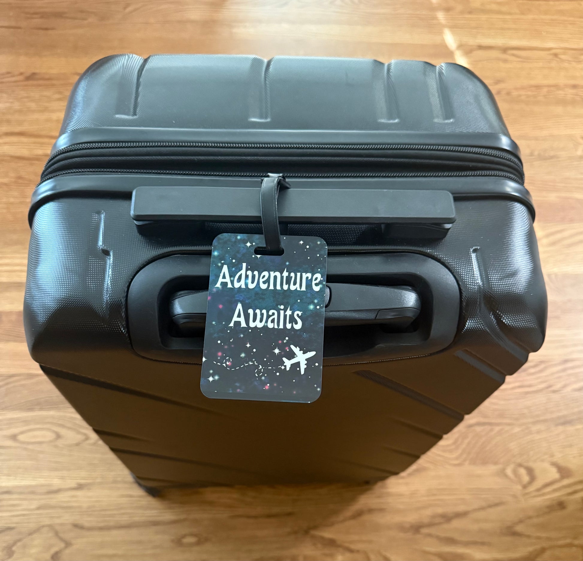 A black luggage tag with the phrase 'Adventure Awaits' written in white, surrounded by star patterns, and an airplane silhouette below Roxzell