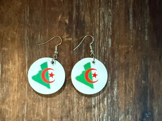 Pair of circular earrings featuring Algeria’s map outline in green with a red crescent and star on a white background, displayed on a wooden surface.