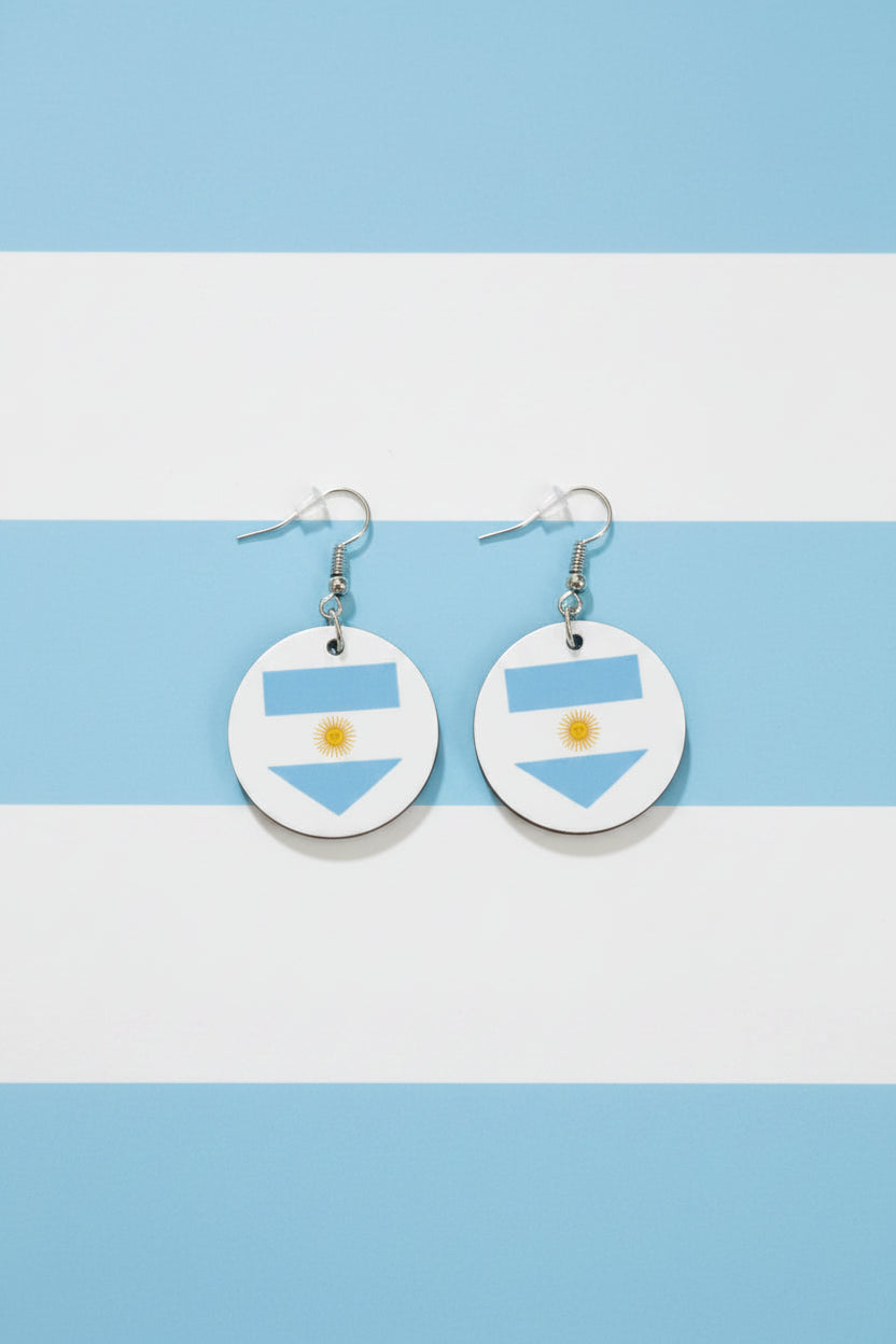 Pair of circular earrings with Argentina’s flag design — two light blue stripes and a white center with a golden sun, displayed on a light blue and white stipe background. Roxzell