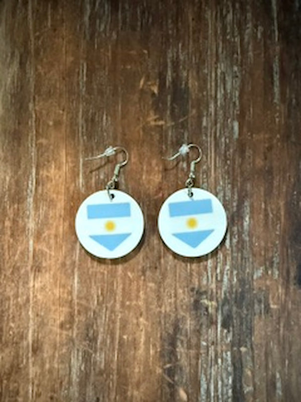 A pair of round drop earrings featuring the light blue and white stripes with a yellow sun emblem of the Argentina flag, hanging from silver fishhook wires on a rustic brown wooden background. Roxzell