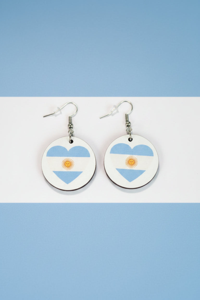 Pair of circular earrings featuring heart shapes in Argentina’s flag colors — light blue and white with a golden sun in the center, displayed on a light blue and white background.