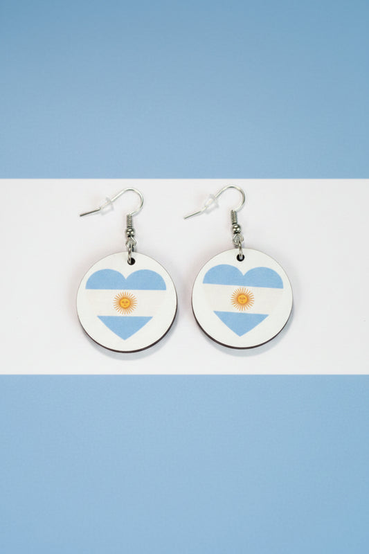 Pair of circular earrings featuring heart shapes in Argentina’s flag colors — light blue and white with a golden sun in the center, displayed on a light blue and white background.