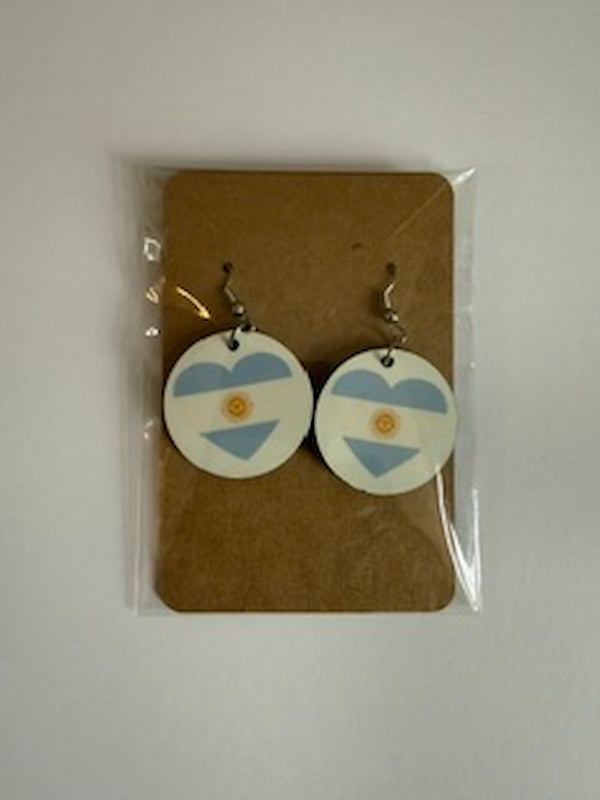 A pair of round drop earrings with an Argentina flag design, displayed on a brown cardboard backing inside a clear plastic sleeve against a light gray background. Roxzell