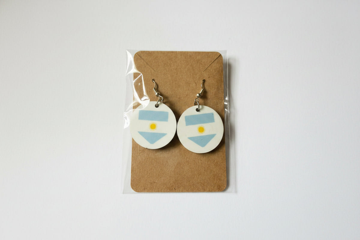 A pair of round drop earrings with silver hooks displayed on a brown cardboard backing inside a clear plastic sleeve. Each earring has an Argentina flag design. Roxzell