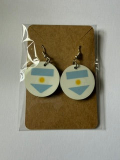Argentina flag earrings attached to a brown backing card inside a clear plastic sleeve, showing light blue and white stripes with a golden sun symbol. Roxzell