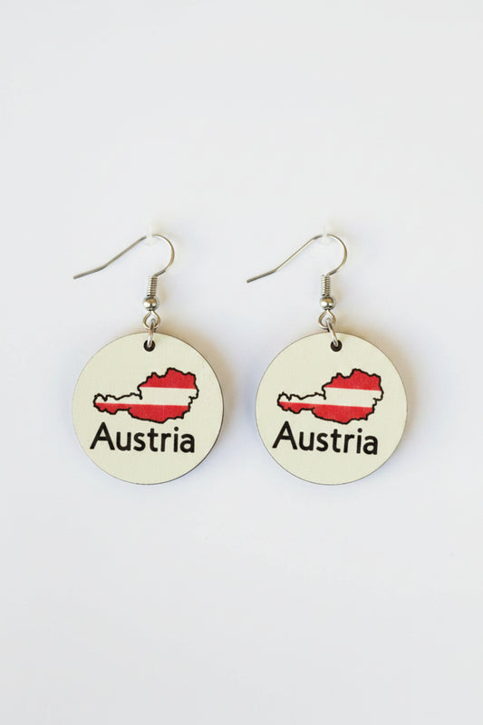 Pair of circular earrings featuring Austria’s map outline filled with red and white flag colors and the word “Austria” printed below, displayed on a white surface. Roxzell
