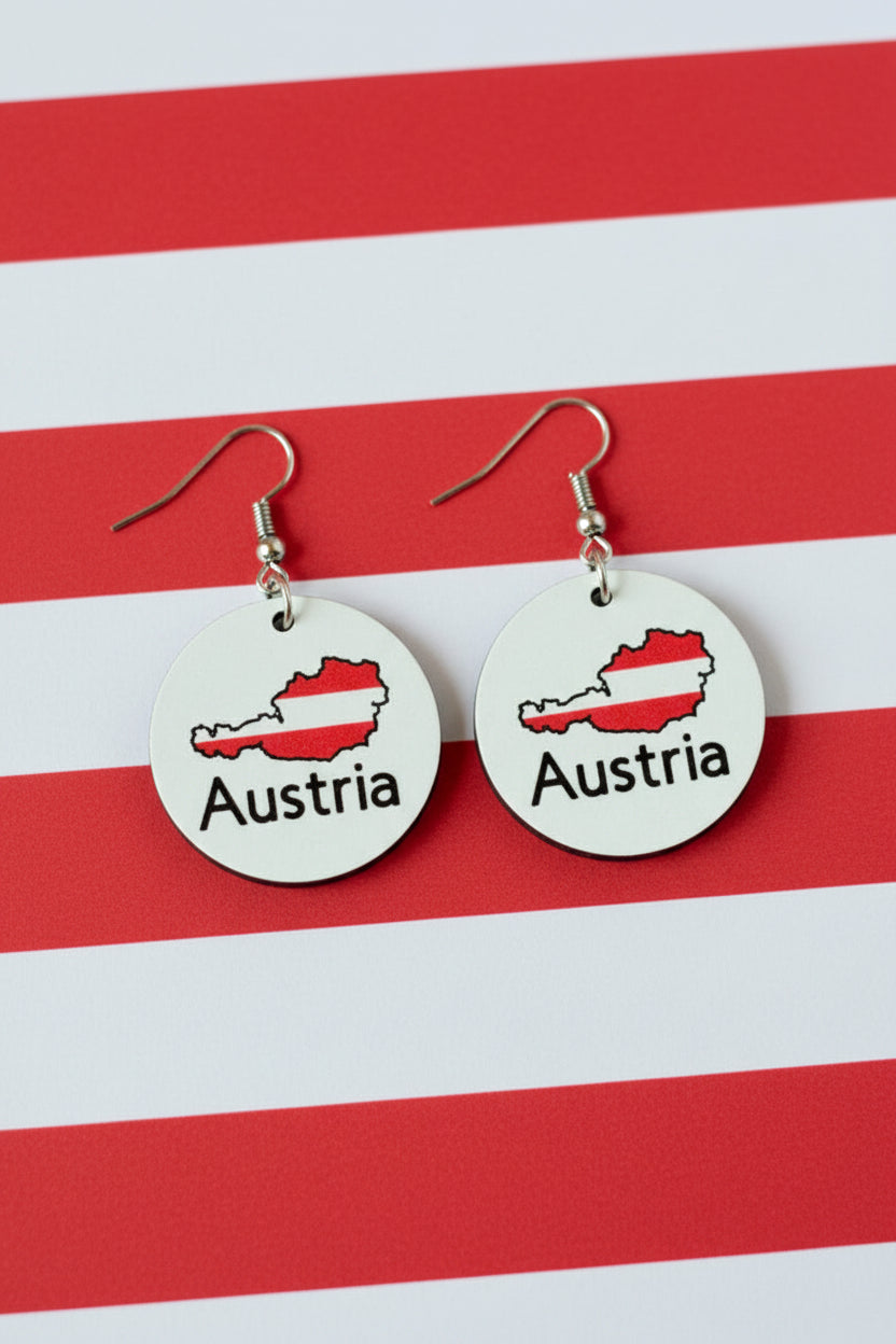 Pair of circular earrings featuring Austria’s map outline filled with red and white flag colors and the word “Austria” printed below, displayed on a red and white surface.