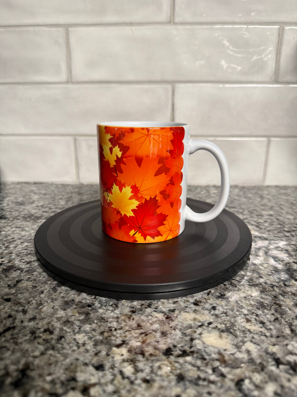 A white ceramic mug with orange, red, and yellow maple leaf pattern on a granite countertop with white tiled backsplash. Roxzell