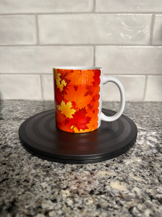 A white ceramic mug with orange, red, and yellow maple leaf pattern on a granite countertop with white tiled backsplash. Roxzell