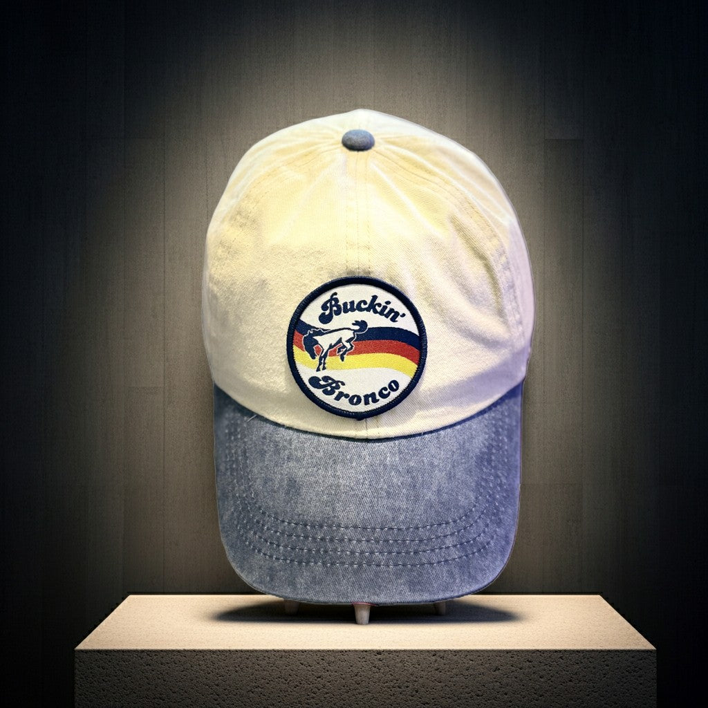 A baseball cap displayed on a pedestal against a dark studio background. The cap has a beige top with a circular embroidered patch reading "Buckin' Bronco," and a blue denim brim. Roxzell