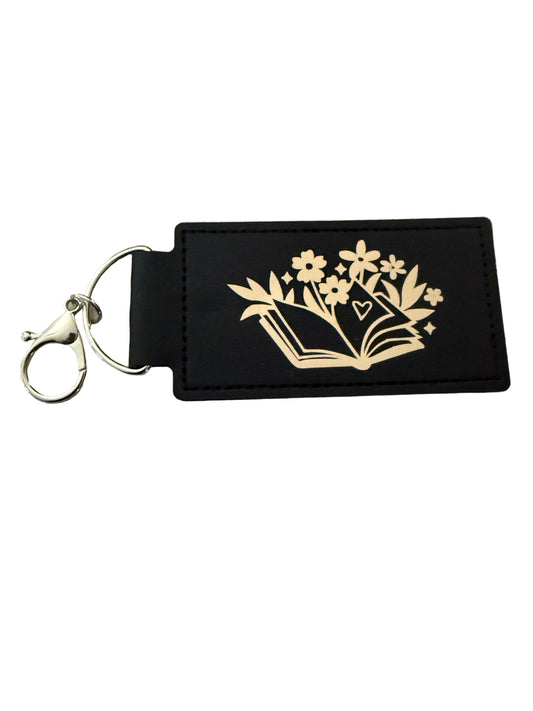 A black faux leather keychain with gold book and floral design, and a gold 'R' initial on reverse side silver d-ring clasp Roxzell