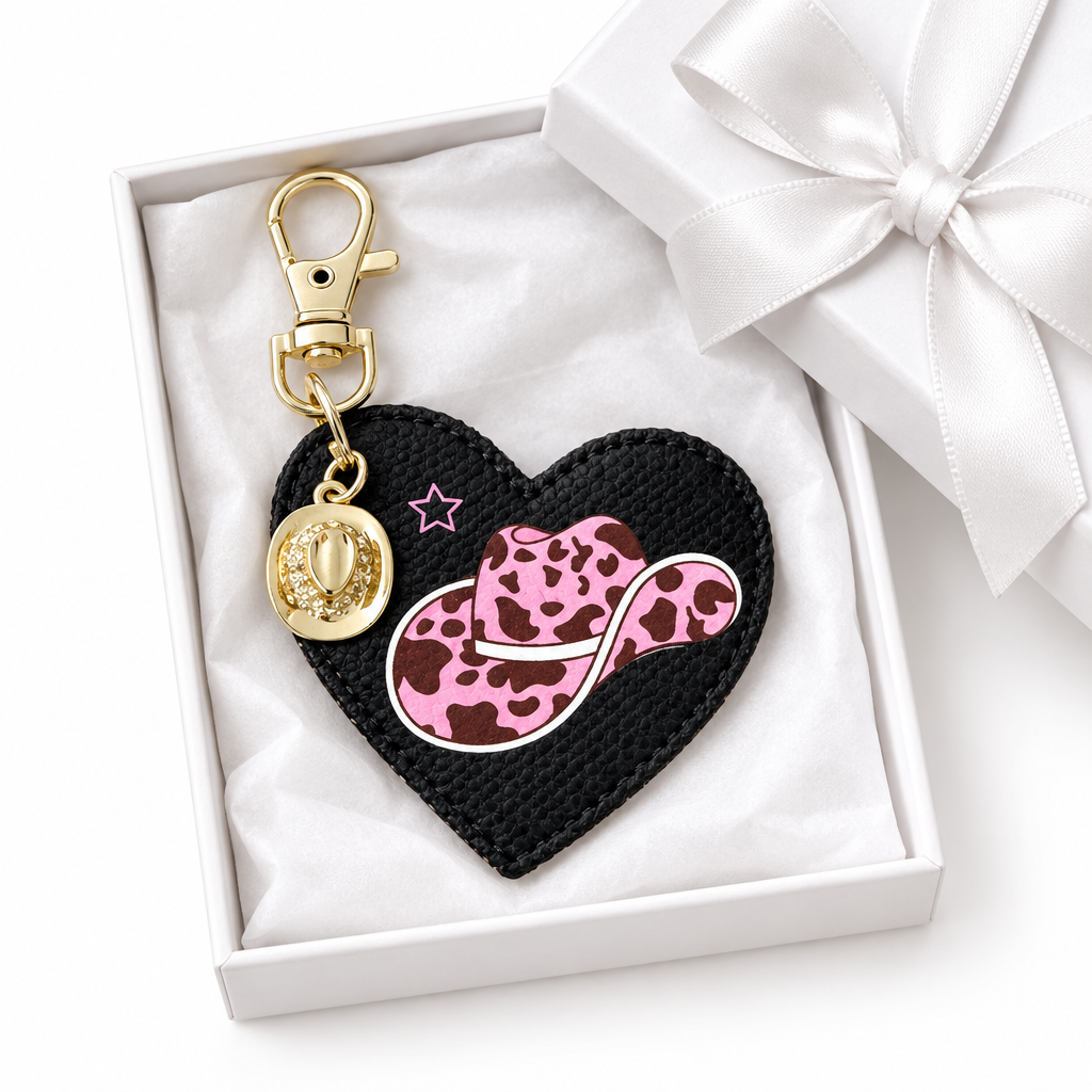 A heart-shaped black keychain charm with a pink leopard-print cowboy hat, gold-toned cowboy hat charm, displayed inside a white gift box with a satin ribbon. Roxzell