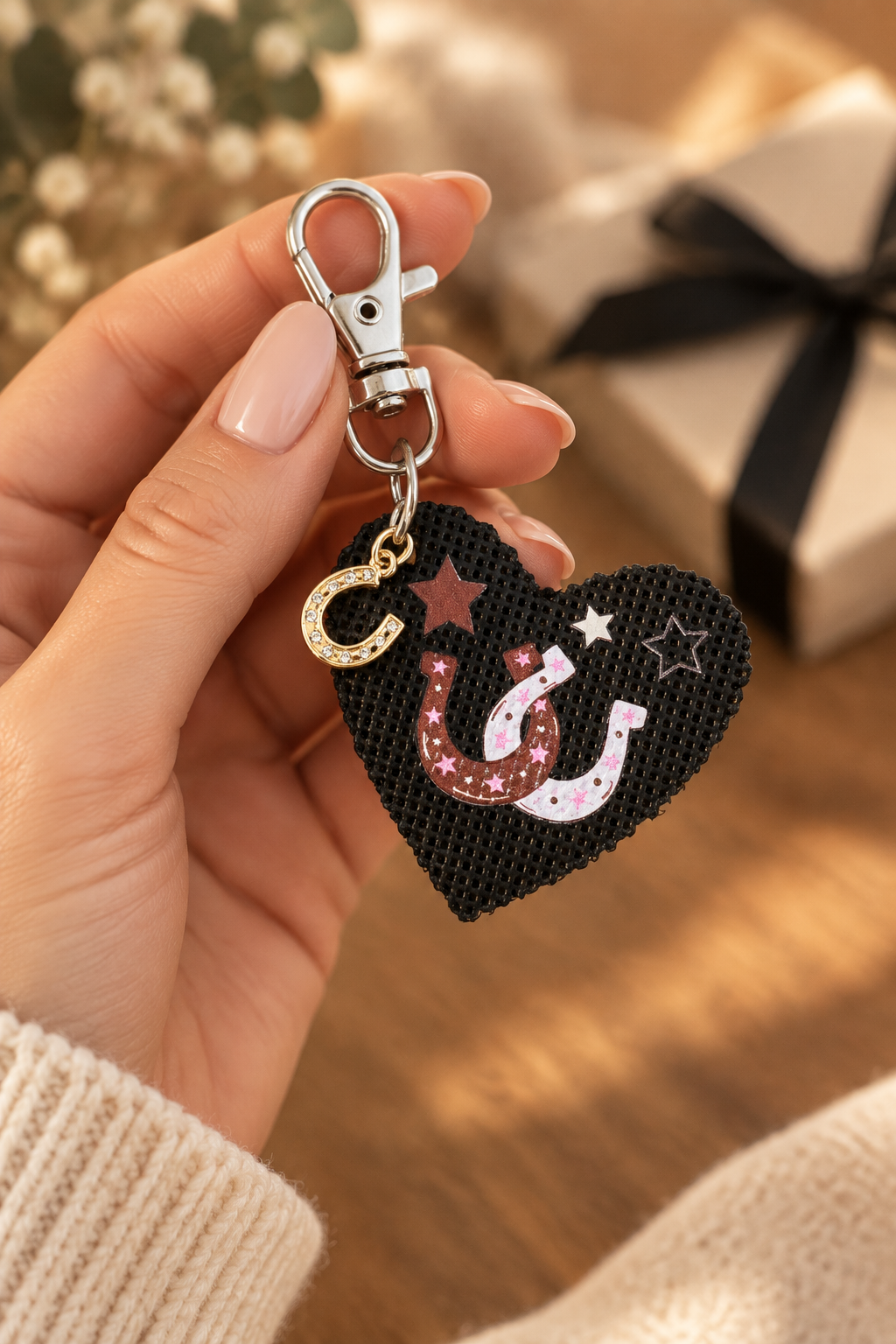 A close-up of a hand holding a heart-shaped black leather keychain charm with horseshoes, stars, and a gold horseshoe charm, attached to a silver clasp. The background is a warm, softly blurred indoor setting with a gift box. Roxzell