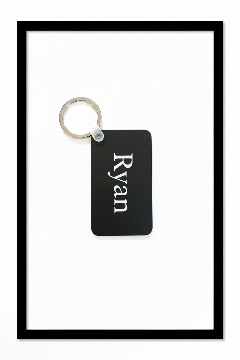 A black rectangular keychain with the name 'Ryan' printed vertically in white serif text, attached to a silver metal split ring, shown on a plain white background with black border. Roxzell