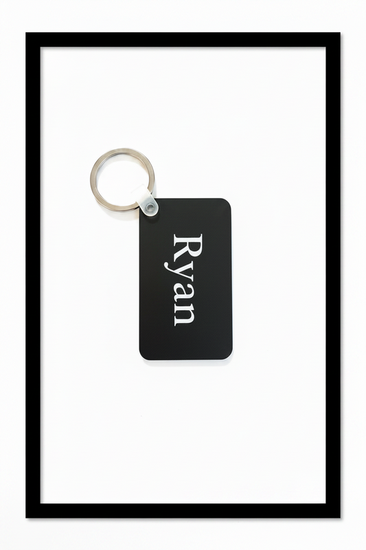A black rectangular keychain with the name 'Ryan' printed vertically in white serif text, attached to a silver metal split ring, shown on a plain white background with black border. Roxzell