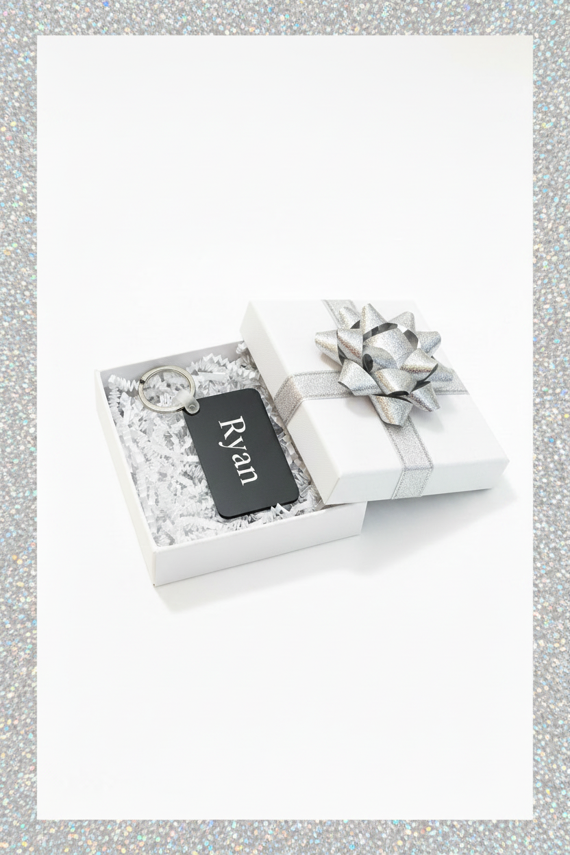 A personalized black metal keychain with the name 'Ryan' in white script, presented inside an open white gift box with white crinkle paper and a silver ribbon bow on a white background and silver border. Roxzell