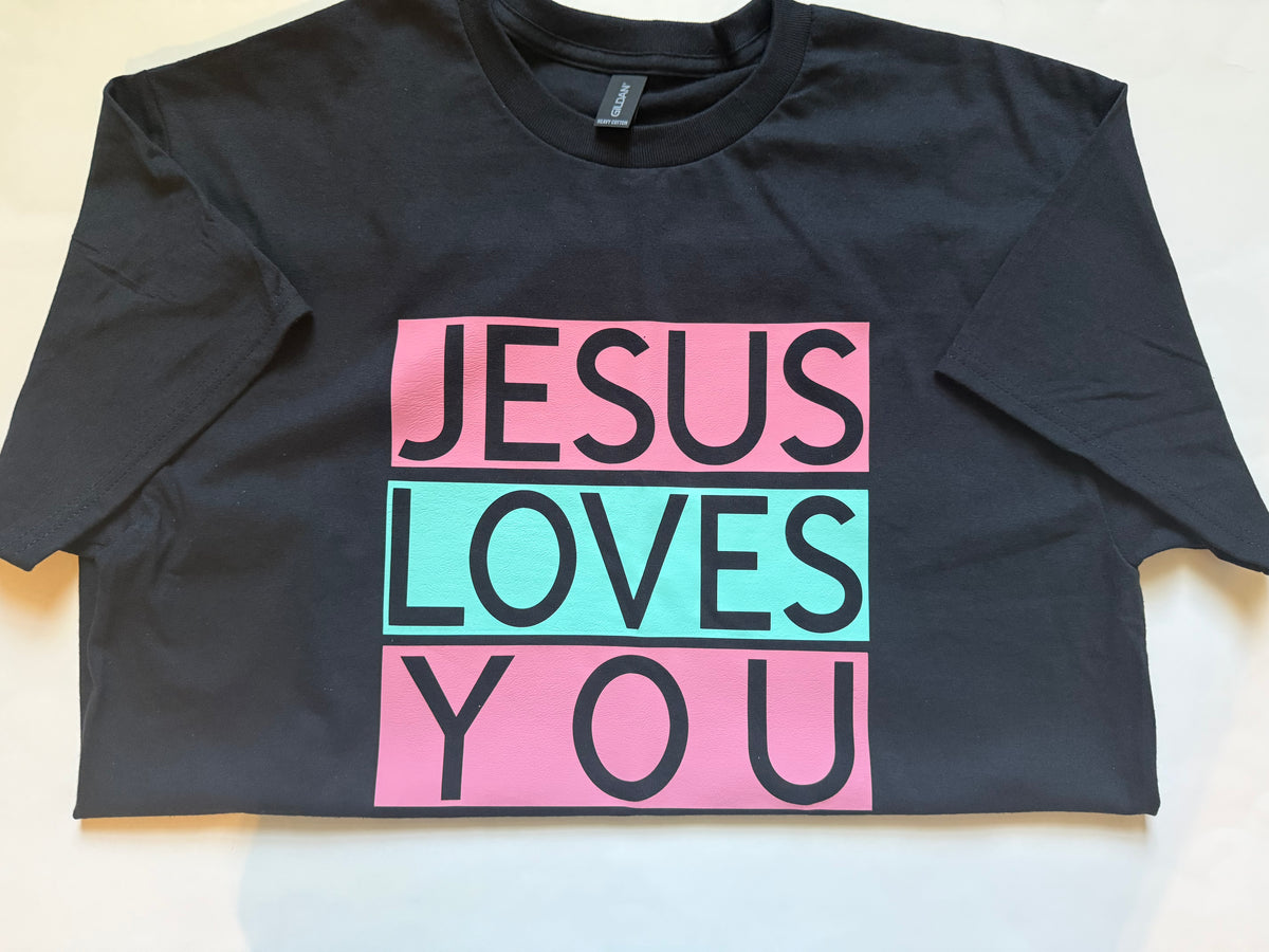 A black short-sleeve t-shirt laid flat on a light surface, featuring a graphic print that reads 'JESUS LOVES YOU' in black text inside pink and green rectangular blocks. Roxzell