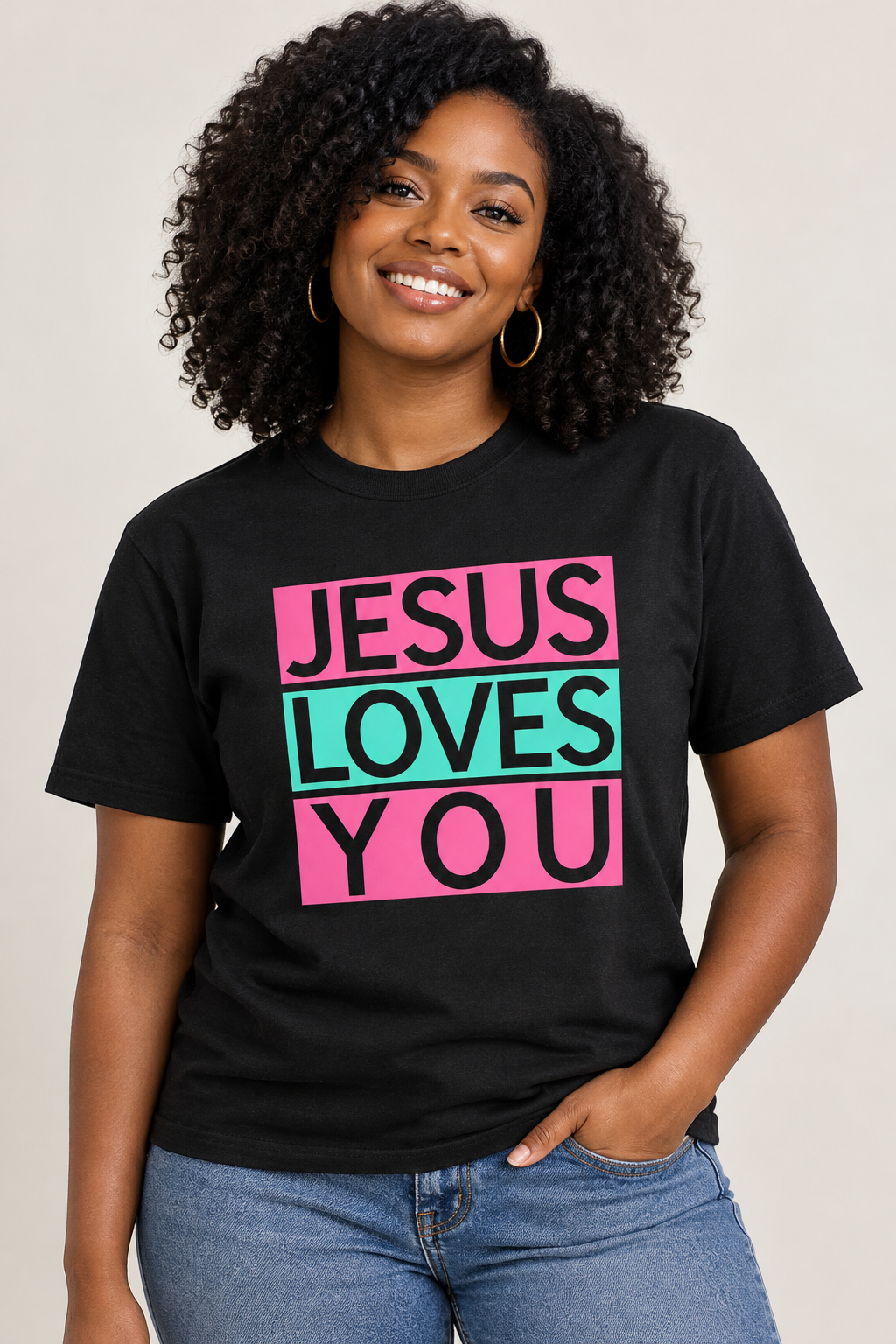 A smiling woman with curly hair wearing a black graphic t-shirt that reads 'JESUS LOVES YOU' in bold pink and teal lettering, paired with blue jeans, against a plain light gray background. Roxzell