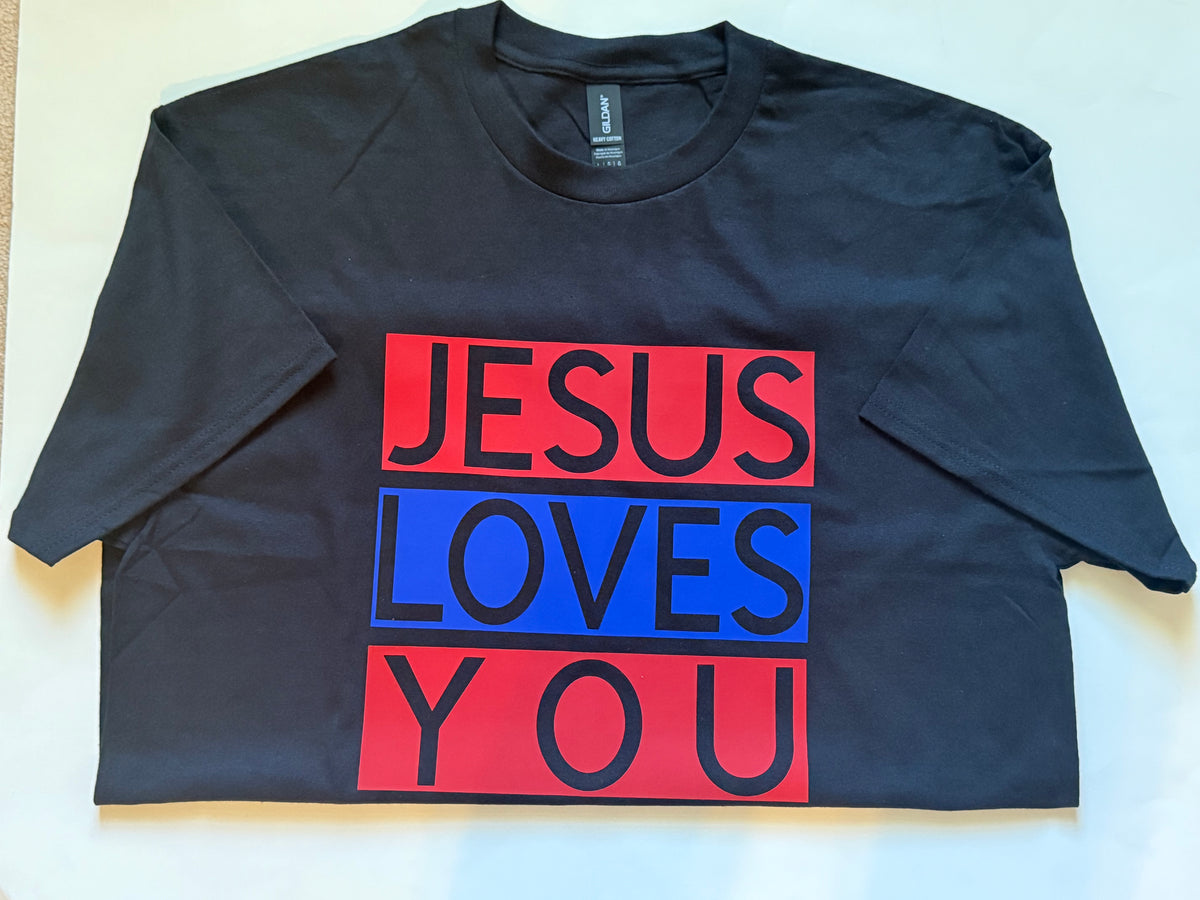 A black short-sleeve t-shirt laid flat on a light background, featuring a graphic print with the words 'JESUS LOVES YOU' in black text inside red and blue rectangular blocks. Roxzell