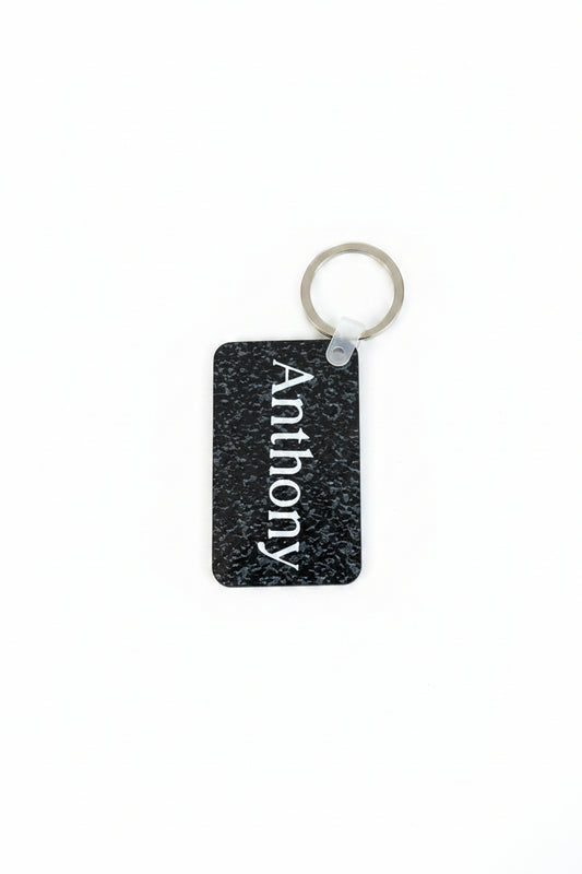 Black rectangular keychain with a speckled texture and the name “Anthony” printed in white, attached to a silver ring on a white background. Roxzell