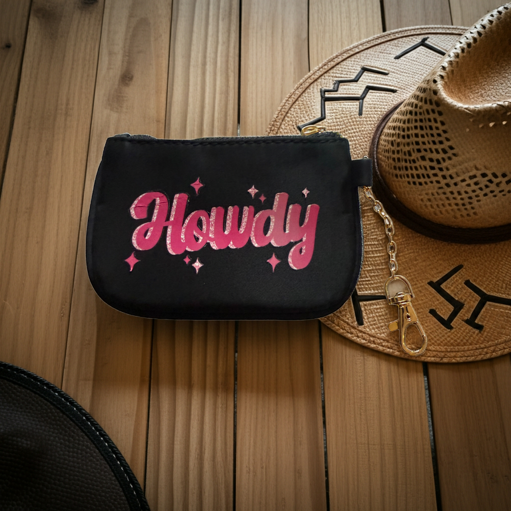 Black id wallet pouch with pink 'Howdy' text on a wooden surface with a cowboy hat.