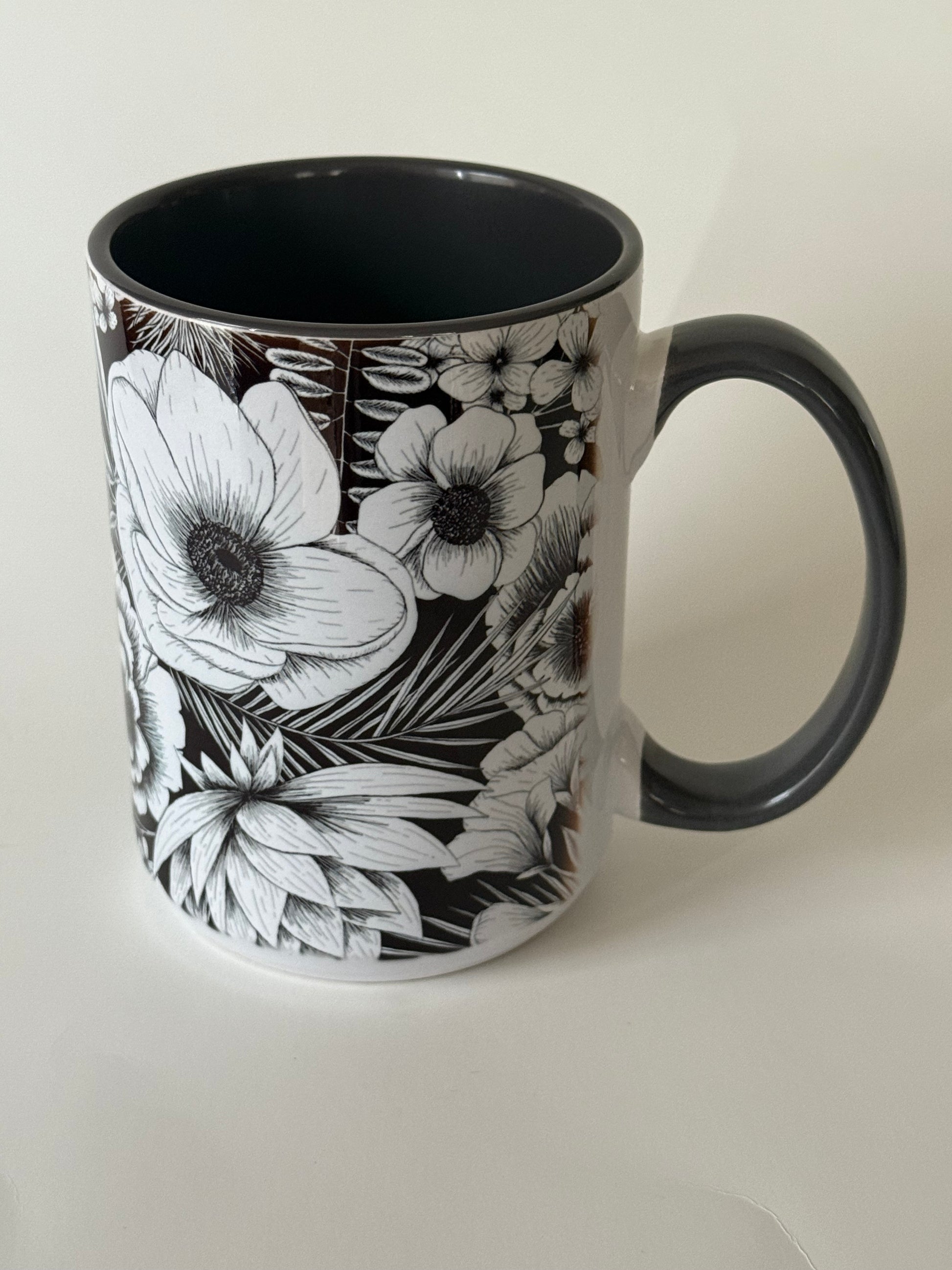A ceramic mug with intricate white botanical flowers on a glossy black background, resting on a white surface. Roxzell
