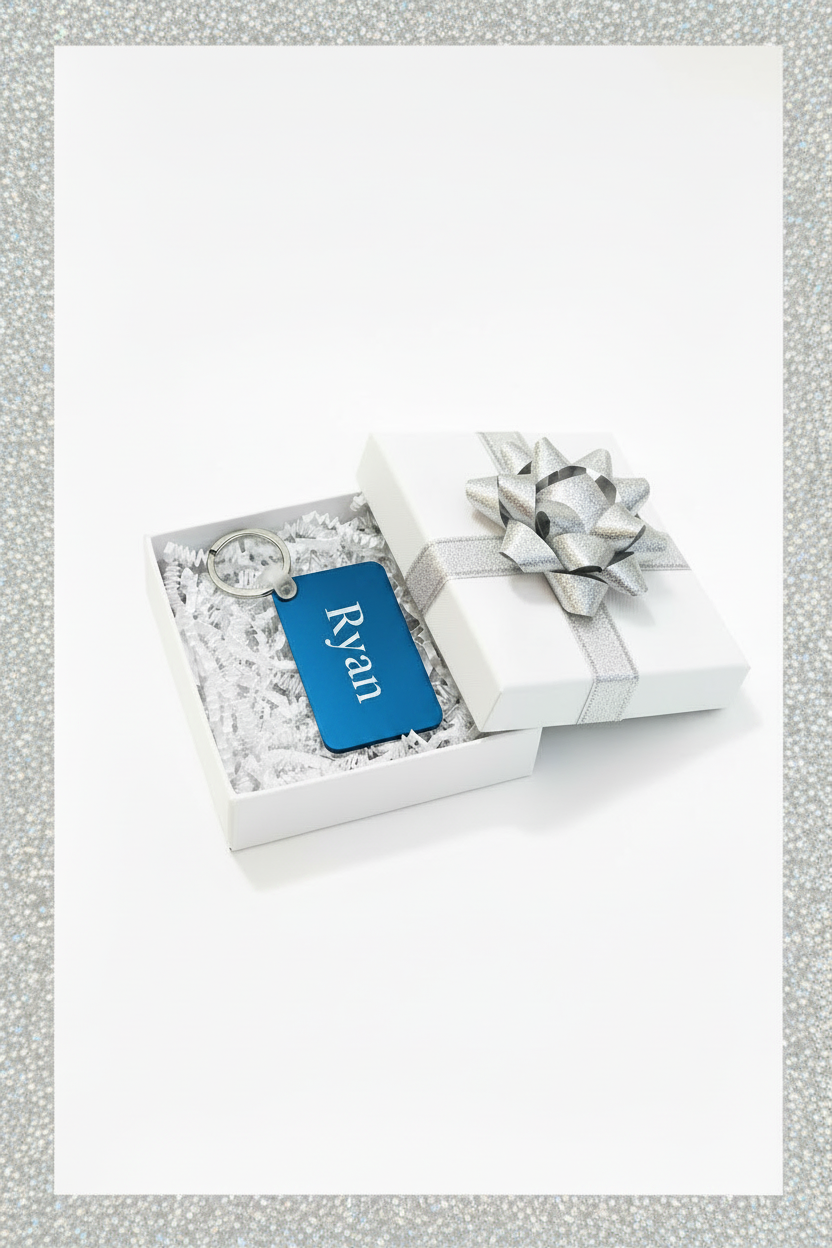 A personalized blue keychain with the name 'Ryan' in white script, presented inside an open white gift box with white crinkle paper and a silver ribbon bow on a white background and silver border. Roxzell