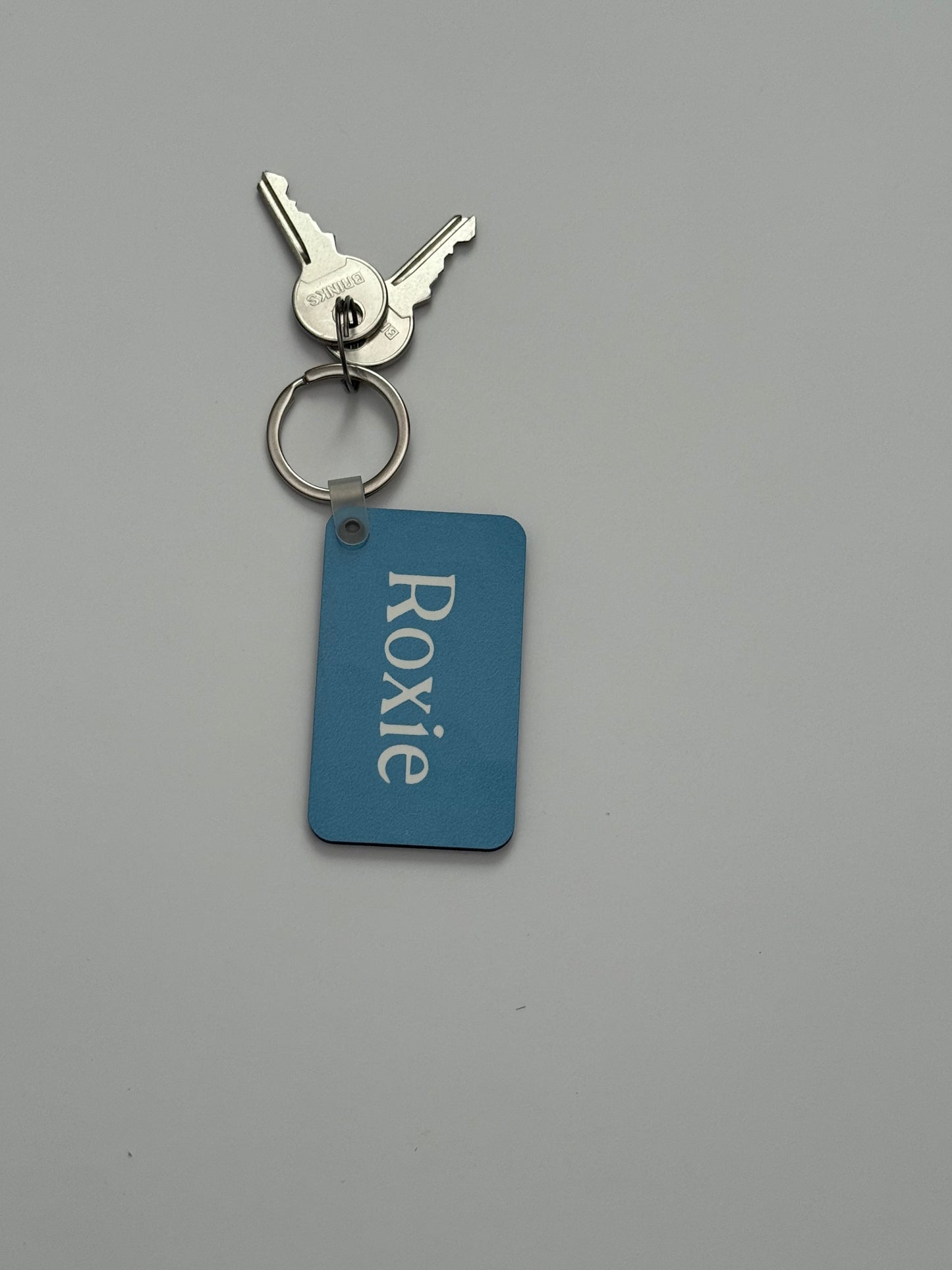 A personalized metal keychain with two silver keys on a split ring and a rectangular blue tag with the name 'Roxie' in white, set against a plain gray background.