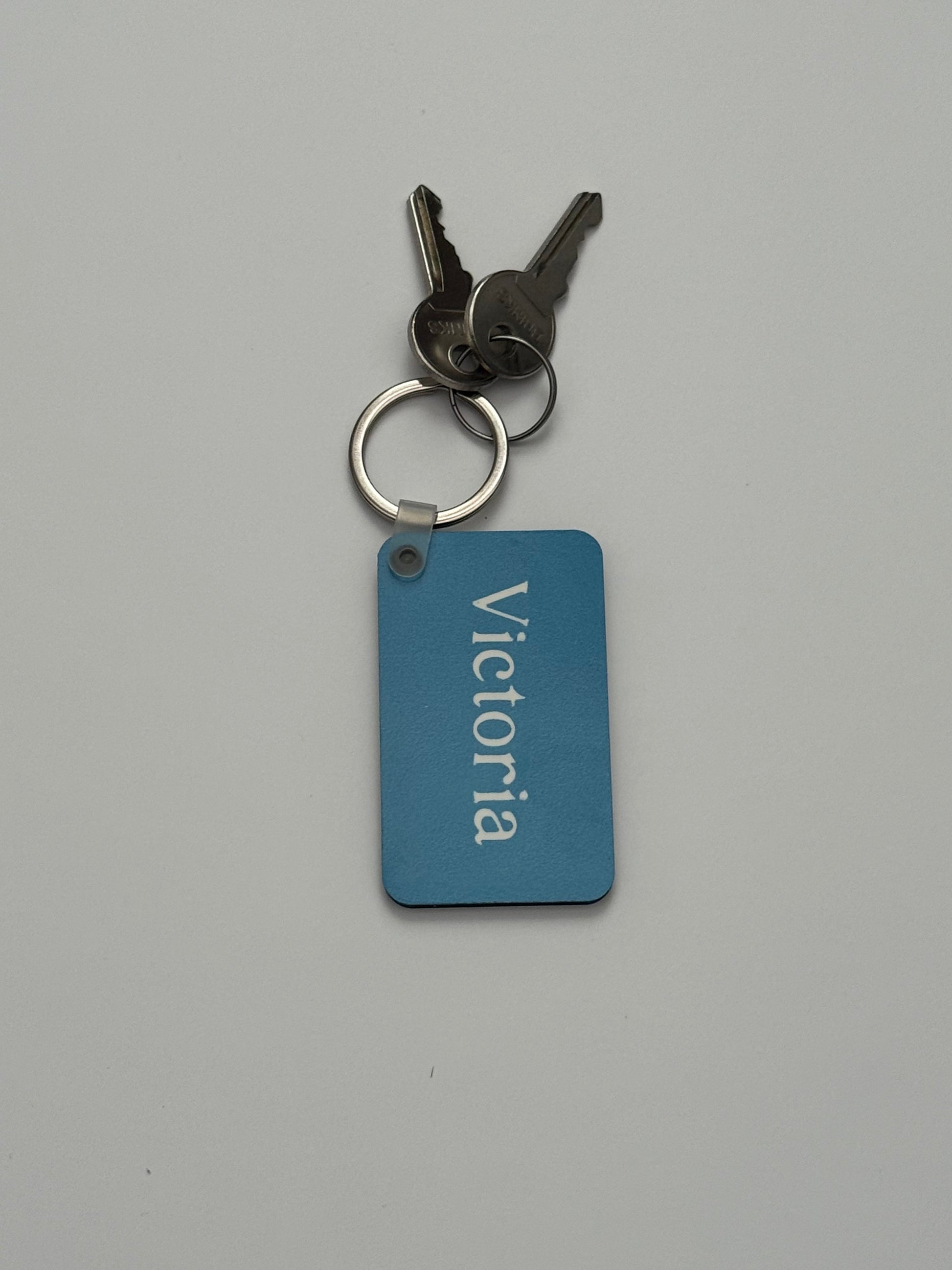 A personalized blue keychain with the name 'Victoria' printed vertically in white, attached to a silver split ring with two keys, on a plain light gray background. Roxzell