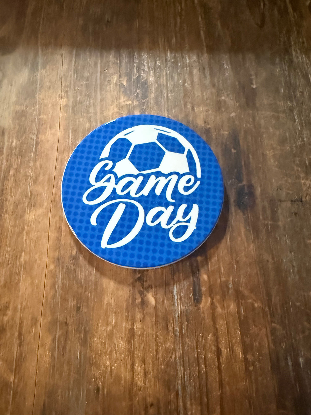 Circular blue coaster with white “It’s Game Day” text, soccer ball graphic, and heart accents on wooden surface. Roxzell