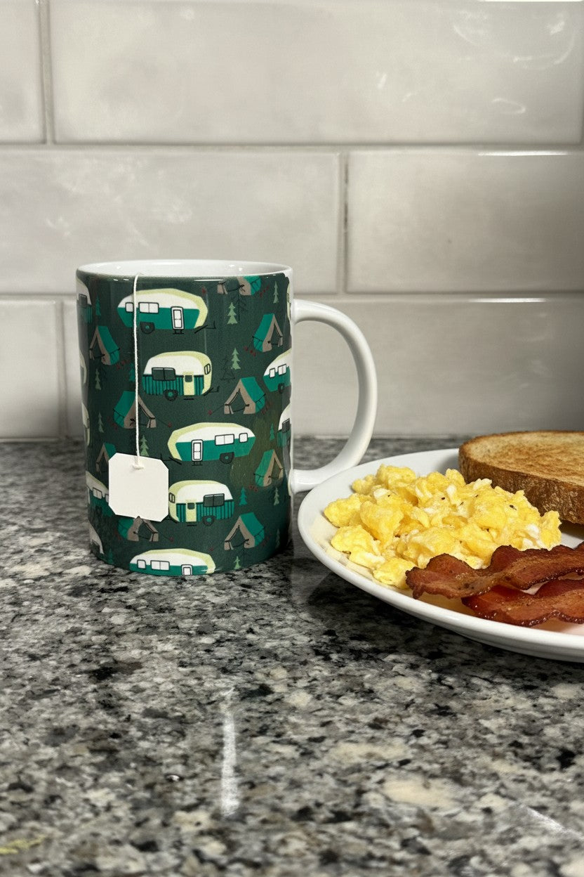 A dark green ceramic mug with a repeating pattern of retro camping trailers and trees, shown on a kitchen counter next to a plate of breakfast with eggs, bacon, and toast.