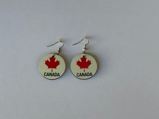 Pair of circular earrings featuring red maple leaves and the word “Canada” printed in black on a white background, displayed on a plain white surface. Roxzell