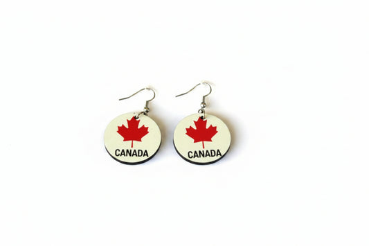Pair of circular earrings featuring red maple leaves and the word “Canada” printed in black on a white background, displayed on a plain white surface. Roxzell