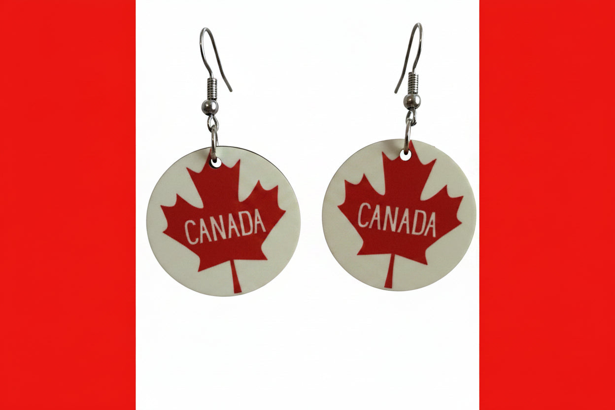 Pair of circular earrings featuring red maple leaves and the word “Canada” printed inside the leaf, displayed on a red and white surface.