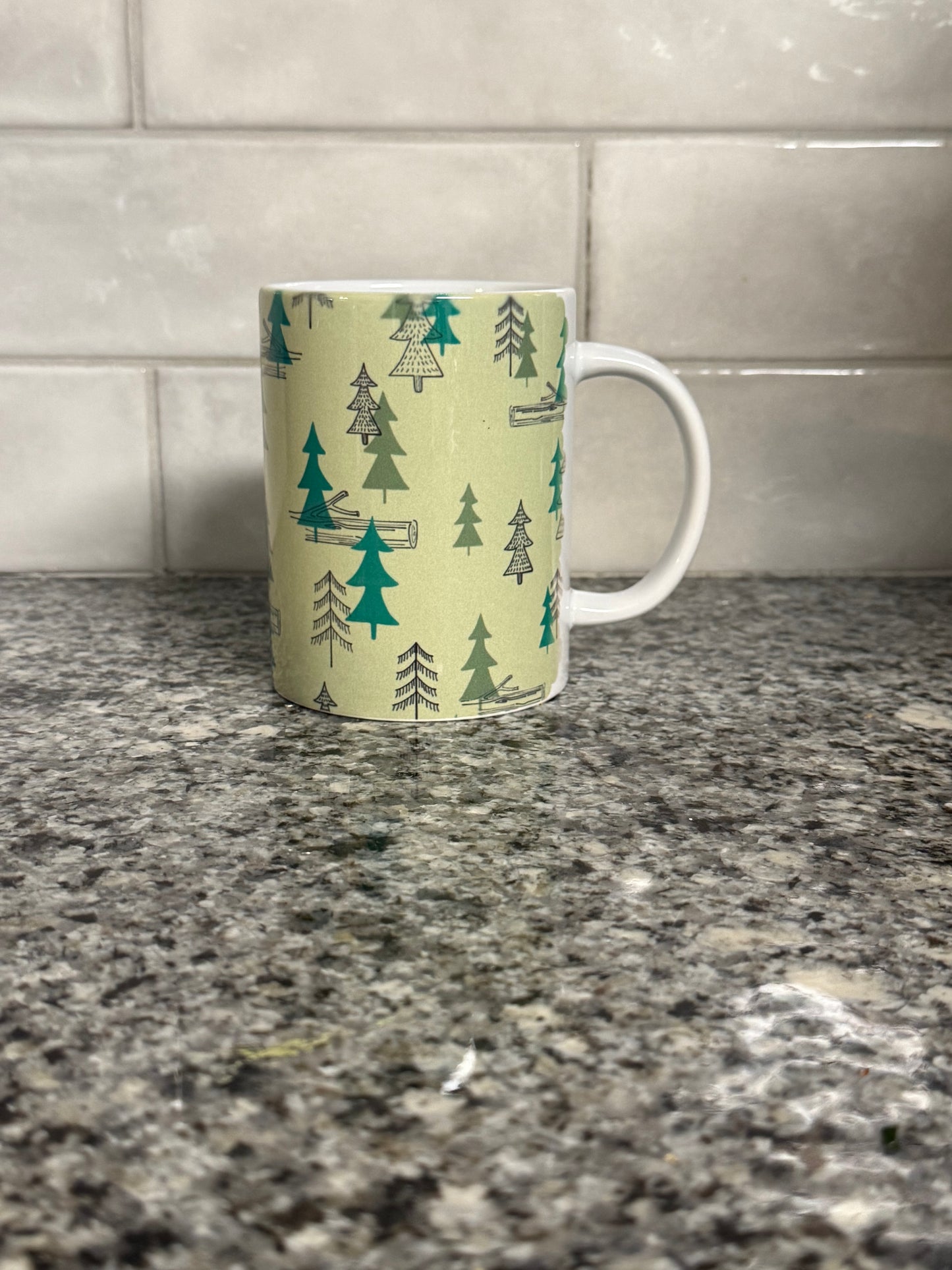 Right side view of a12oz coffee mug with camping site forest trees design on a granite countertop. Roxzell