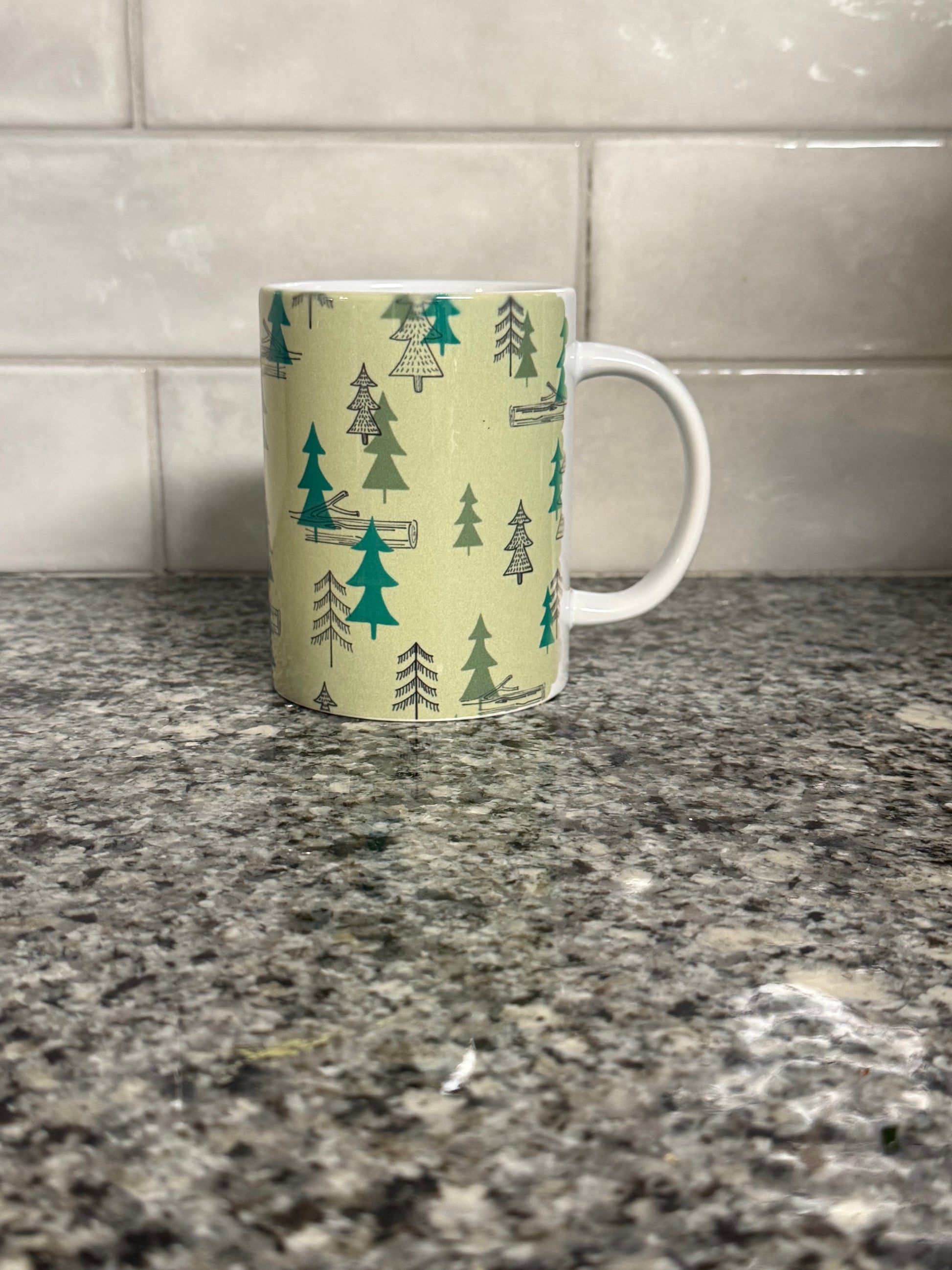 Right side view of a12oz coffee mug with camping site forest trees design on a granite countertop. Roxzell