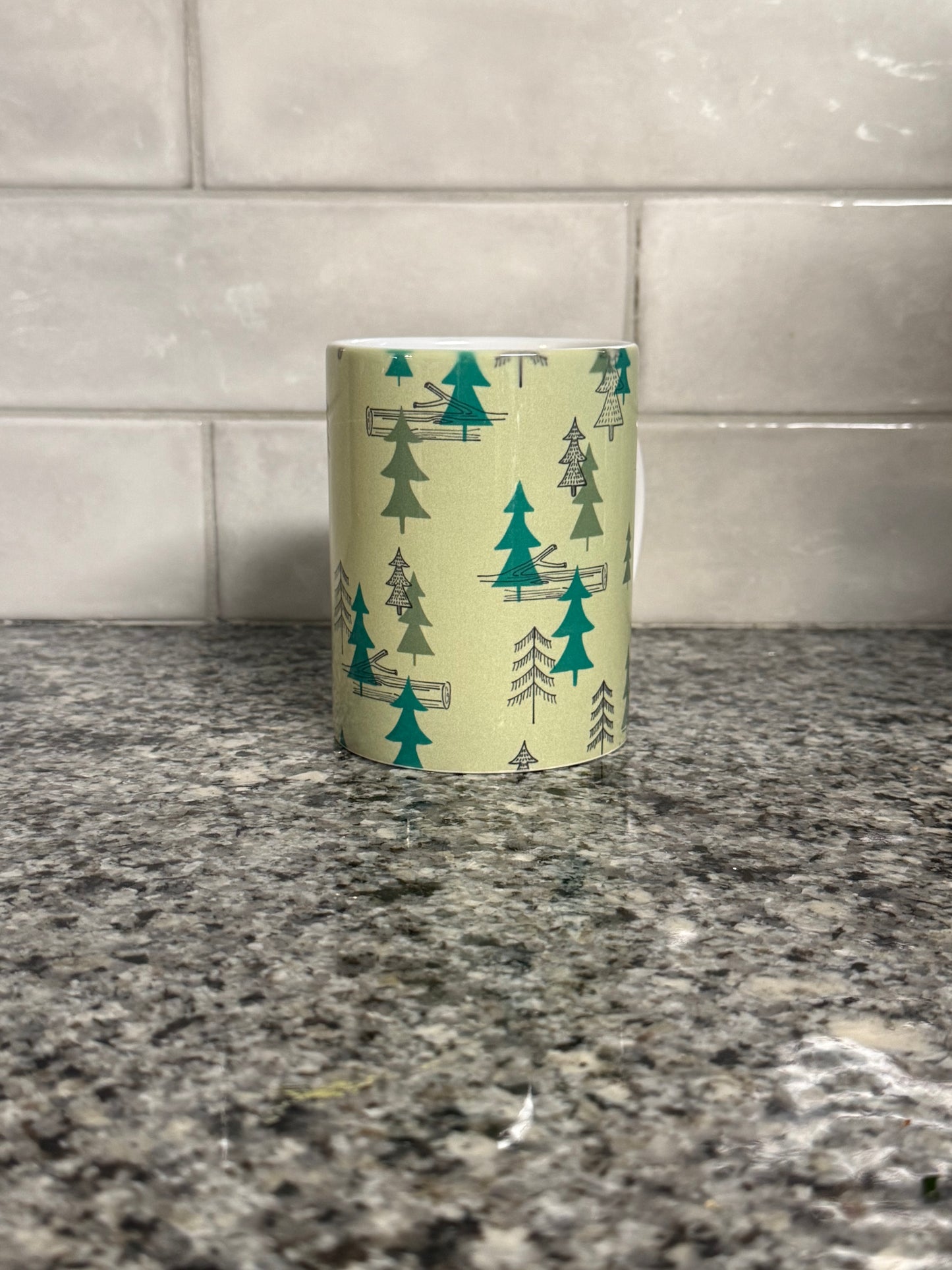 Center view of a 12 oz coffee mug with camping site forest trees design on a granite countertop. Roxzell
